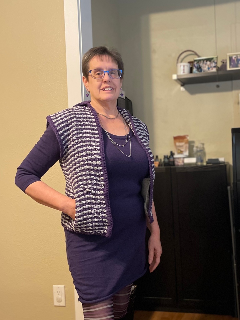 A white woman with short brown hair and blue glasses models a gray and purple crocheted vest over a purple, long-sleeved dress with her right hand in the pocket of the vest.